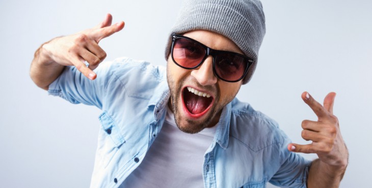 Cool and funky. Top view of handsome young man in hat and sunglasses gesturing and grimacing while standing against grey background