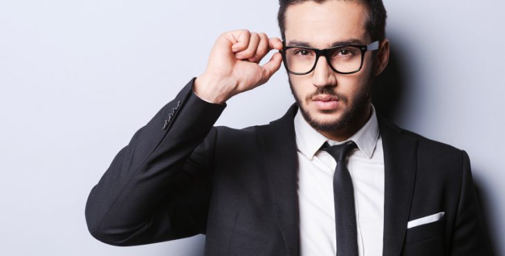 Taking life seriously. Portrait of handsome young man in formalwear adjusting his glasses while standing against grey background