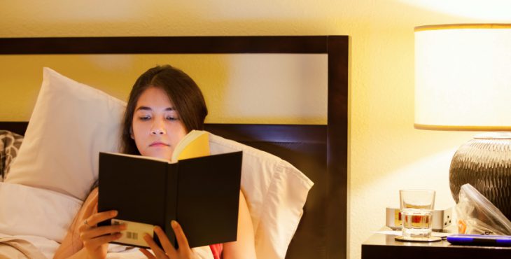 Biracial teen girl reading book  in bed at night